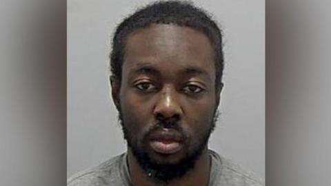 Tyreese Kamau: A man with black hair and a black beard wearing a grey and round necked t-shirt in his custody photo