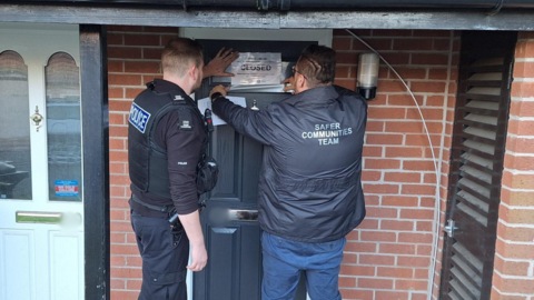 A police officer and member of the Safer Communities Team place a sign which reads "closed" on a dark grey front door surrounded by red brick.