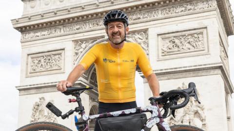 Chris Boardman standing behind his bike in front of the Arc de Triomphe, in Paris. He is wearing a bike helmet and a yellow top.