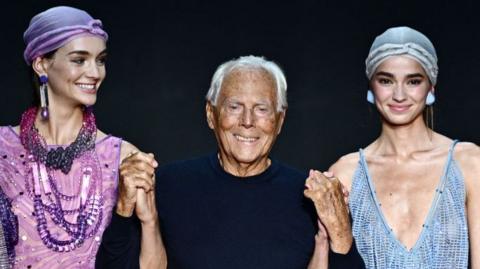 A photohraph of Giorgio Armani standing in the middle with a model standing to his left and a model standing to his right 