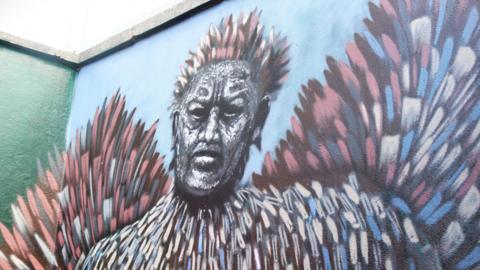 A vibrant mural fills the corner of an outdoor wall, showing a stylized human figure whose face is intentionally blurred or abstracted. From the figure’s head and shoulders, large feather‑like shapes burst outward in sweeping arcs of pink, blue, white, and brown, giving the impression of motion, energy, or transformation.