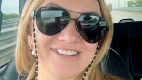 Polina Azarnykh looking straight at the camera while seated in a vehicle. She has long blonde hair and is wearing large sunglasses and a black top and is smiling.