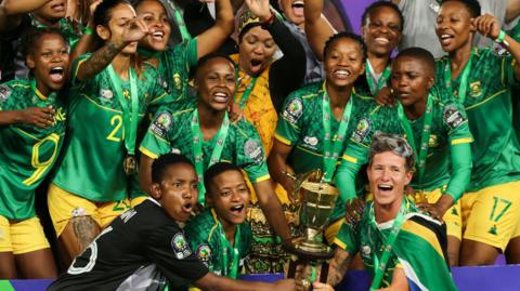 A jubilant South Africa team, some with medals around their necks on green ribbons, pose with the Women's Africa Cup of Nations trophy in 2022