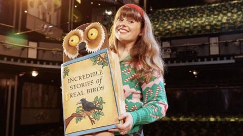 Georgie Samuels in her costume in the West End show, wearing a green top with candy canes and holding a big book with an owl picture saying "Incredible Stories of Real Birds"