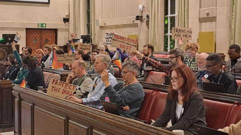 A small crowd of people sat in a public gallery at a council meeting. Many people are holding up banners and flags. Some of the banners say "protect the dolls", "trans men are men" and "trans people exist". Nobody is looking at the camera.
