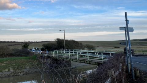 The single lane bridge with the river running underneath it. Cars can be seen on the far side and in the distance you can see the rolling hills of the South Downs.