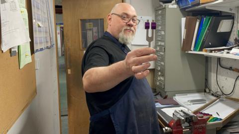 A person stands in a workshop holding a metal tuning fork with purple weights near the tips. The workspace includes tools, drawers, a vice labelled “RECORD 74,” and neatly arranged metal pieces on a bench. Behind the person are a corkboard with papers, shelves with folders, and an open door leading to another room.