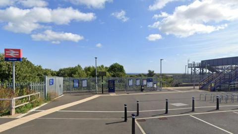 Horden train station: a sign with a white rail logo on a red background, with Horden written underneath, stands in a parking area situated next to rail tracks and a rail bridge.