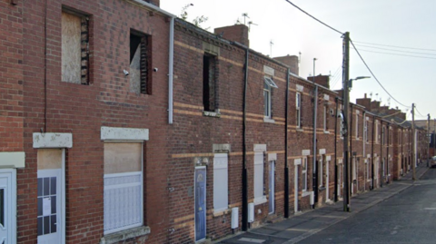 A street of vandalised terraced homes with boards up in the windows and pictures of fake front doors stapled on to the fronts of boards over the front doors.