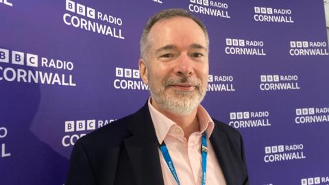 Close-up of Royal Cornwall Hospitals Trust CEO Steve Williamson who has dark hair and a beard. He is standing in front of a purple BBC Radio Cornwall backdrop. Mr Williamson is wearing a pink shirt and a black jacket and has a blue Royal Cornwall Hospitals Trust lanyard around his neck. 