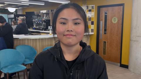 A teenage girl with long black hair tied back, wearing a black jacket, stands in a cafe