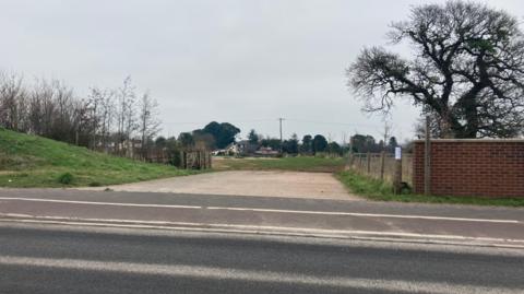 The entrance to a field. There is a tree and a brick wall to the right.