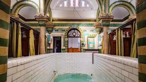 A highly decorated room with green and mustard tiles and ornate columns. There is a deep plunge pool in the foreground with white tiles. At the sides there are booths separated by wooden panels with decorations at the top and green privacy curtains.