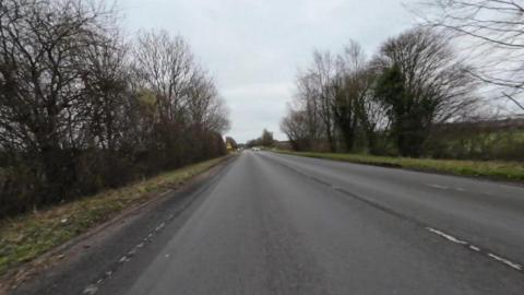 A shot of the road from the perspective of a car travelling down the road. There are trees and grass at the sides of the road and a grey sky, as well as cars and a road sign in the distance. 