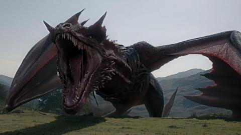 CGI of a giant red-black dragon, open mouthed, its wings out-stretched as it appears to stride rather than fly atop a mountainous landscape