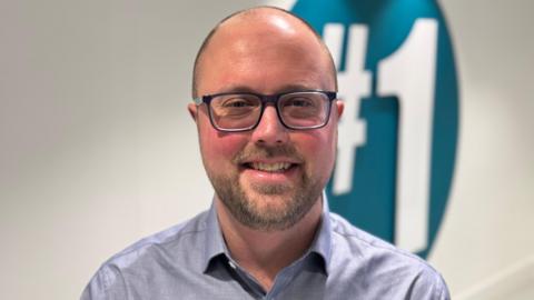 Olly Newton smiling. He is wearing a blue shirt and dark rimmed glasses. He has short hair and a beard.