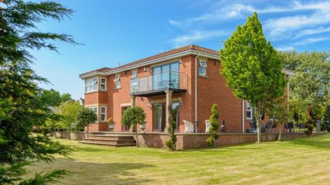 Red Gables. It is a two-storey brown-brick house with a balcony on the second floor and a seating area outside. It is surrounded by trees and other plants.