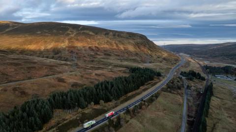 An aerial view of the A9 at Glen Garry. There are two lorries, both white, one with a green trailer and the other with a red trailer, on the road in the foreground. In the background, greenery including trees and grass can be seen going up the side of a hill, which is partially in the sun. The sky is blue and grey.