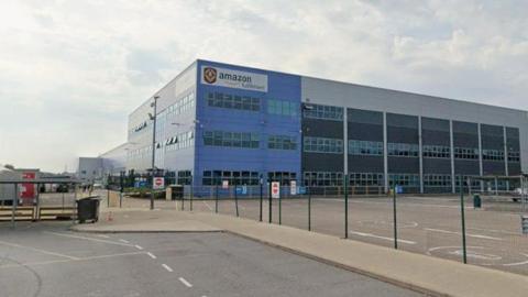A large warehouse building, viewed from a car park. The corner of the building has blue cladding and an Amazon company logo on a white background at the top.