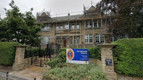 The entrance of Wolsingham School. It is a two-storey stone building which was covered in scaffolding when the picture was taken. It is surrounded by trees and shrubs. There is a blue sign on the fence with the school logo, reading Wolsingham School.