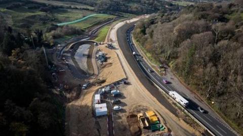 An aerial view of a new stretch of road, which is still under construction, beside an existing road being used by traffic up a hill between two woodlands.