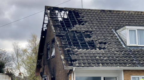 The roof of a bungalow is shown, charred by the fire to the point where there are gaps in the roof. The whole house isn't burned, and some trees can be seen in the background.