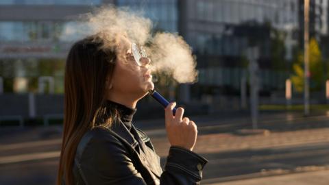 A woman with long brown hair and glasses is smoking a disposable vape. There is a puff of white smoke in the air. She is wearing a black jacket and turtle neck as she stands on the street.