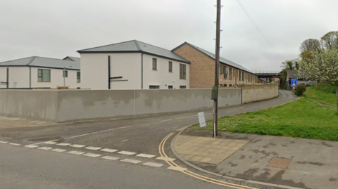 The turning in to Foundry Lane. One side has houses and the other is a grassy verge and a telegraph pole. At the furthest visible point of the lane there is a railway bridge.