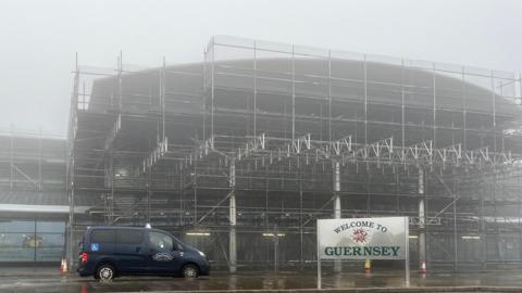 Guernsey airport with fog. A blue taxi is parked at the front. There is also a sign saying welcome to Guernsey. The airport building is covered in scaffolding,