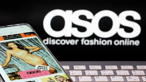 A phone displaying the Asos app, with a female model showing off a gold dress, is seen against a backdrop of the Asos logo on a computer screen with some of the keys on the keyboard in view, taken in 2021