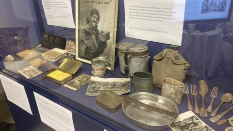 An exhibition display of old utensils including discoloured metal spoons and forks. There are various rusted and battered tins and pots with handles, along with a WW2 poster depicting a man offering rum.