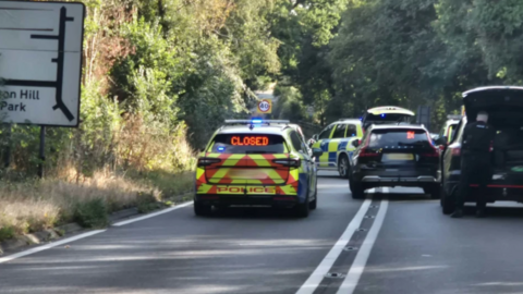 Police cars can be seen in the road with other unmarked vehicles blocking the route. There is a road sign on the left with trees lining the road.