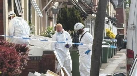 Officials in white protective overalls stand at the door of a building. In the background are other houses with green bins lined up outside and a vehicle on the right.
