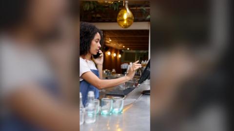 A woman wearing an apron behind a bar holds a phone to her ear with one hand and is typing on a screen with her other