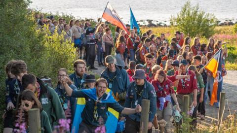 A long line of scouts, some with flags, makes its way around a bend near the sea