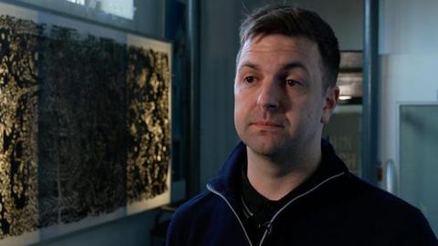 Conchúr Ó Muadaigh - a man with short brown hair, wearing a navy jumper, stood beside a large framed print