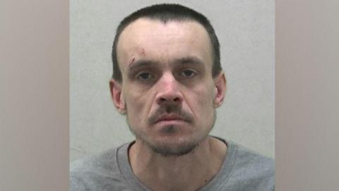 Mugshot of Craig Howse who is wearing a grey sweatshirt. He has very short dark hair and slight beard and moustache and has a cut above his right eye. 