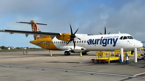 An Aurigny aircraft with its yellow and white livery. It has two propellers. The aircraft is on a runway at an airport.