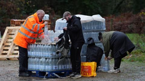 A person in an orange high-vis jacket handling a stack of bottled water in a car park with two members of the pubic loading bottles into bags.