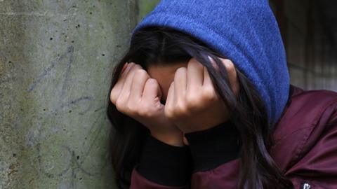 Generic image of a teenage girl with long dark hair with her head in her hands. She is leaning against a graffitied wall and is wearing a blue hoodie under a burgundy bomber jacket