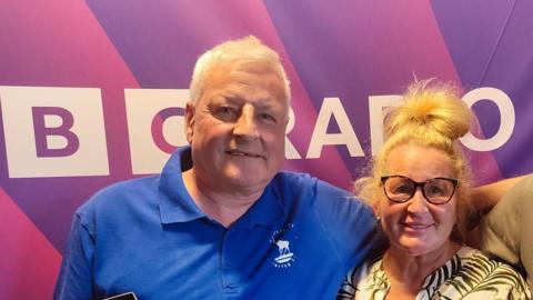 Charlie and Alison are posing in front of a 91ȱ Radio Tees sign. He is wearing a blur polo shirt and has short white hair. She is wearing a green  and white top and has blonde hair and brown glasses.