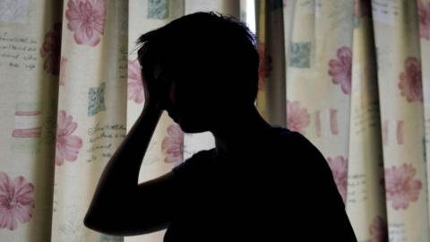 A silhouette of a person with their hand on their head, behind them are curtains with pink flowers on which are closed across a window