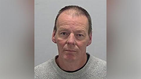 A mugshot of a man with short black hair wearing a grey jumper with a black top underneath.