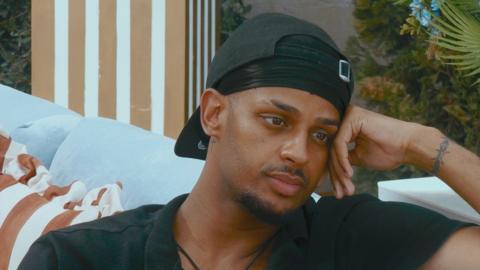A man wearing a backwards cap rests his head on his hand while sitting on a couch.