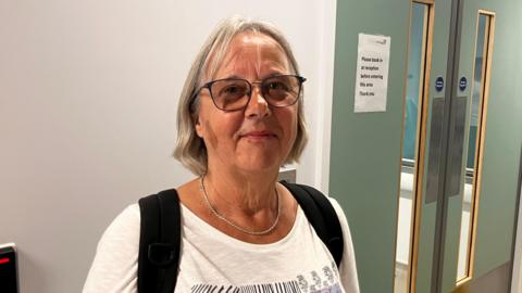 Renate Kennedy in A&E in Bournemouth. She has shoulder length blonde hair and is wearing a white top with the black straps of a bag over her shoulders. She is wearing glasses and a silver necklace. She is standing in front of a white wall with green double doors.