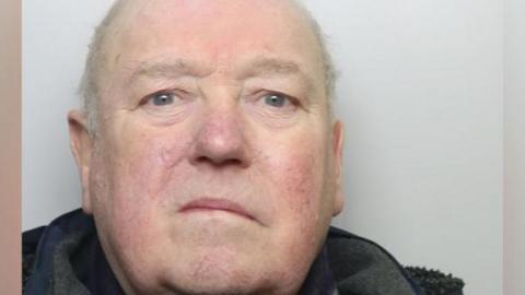 A police mugshot of a man in his eighties, mainly bald but with wispy white hair. He is wearing a navy coat and staring directly into the camera.