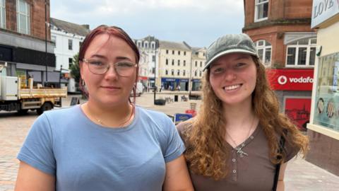 A woman with a blue t-shirt and silver glasses stands next to a woman with a grey cap and brown t-shirt