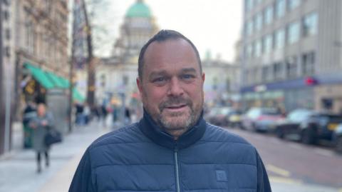 A man in a navy winter jacket looks at the camera. He has short dark hair and a dark short beard. Behind him is Belfast city hall and the high street. 