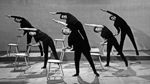 A black and white image of people dressed in black doing exercises in the studio using chairs.