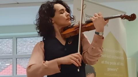 Woman with dark curly hair playing a violin, she has her eyes closed and wears a dark waistcoat and dusky pink shirt.